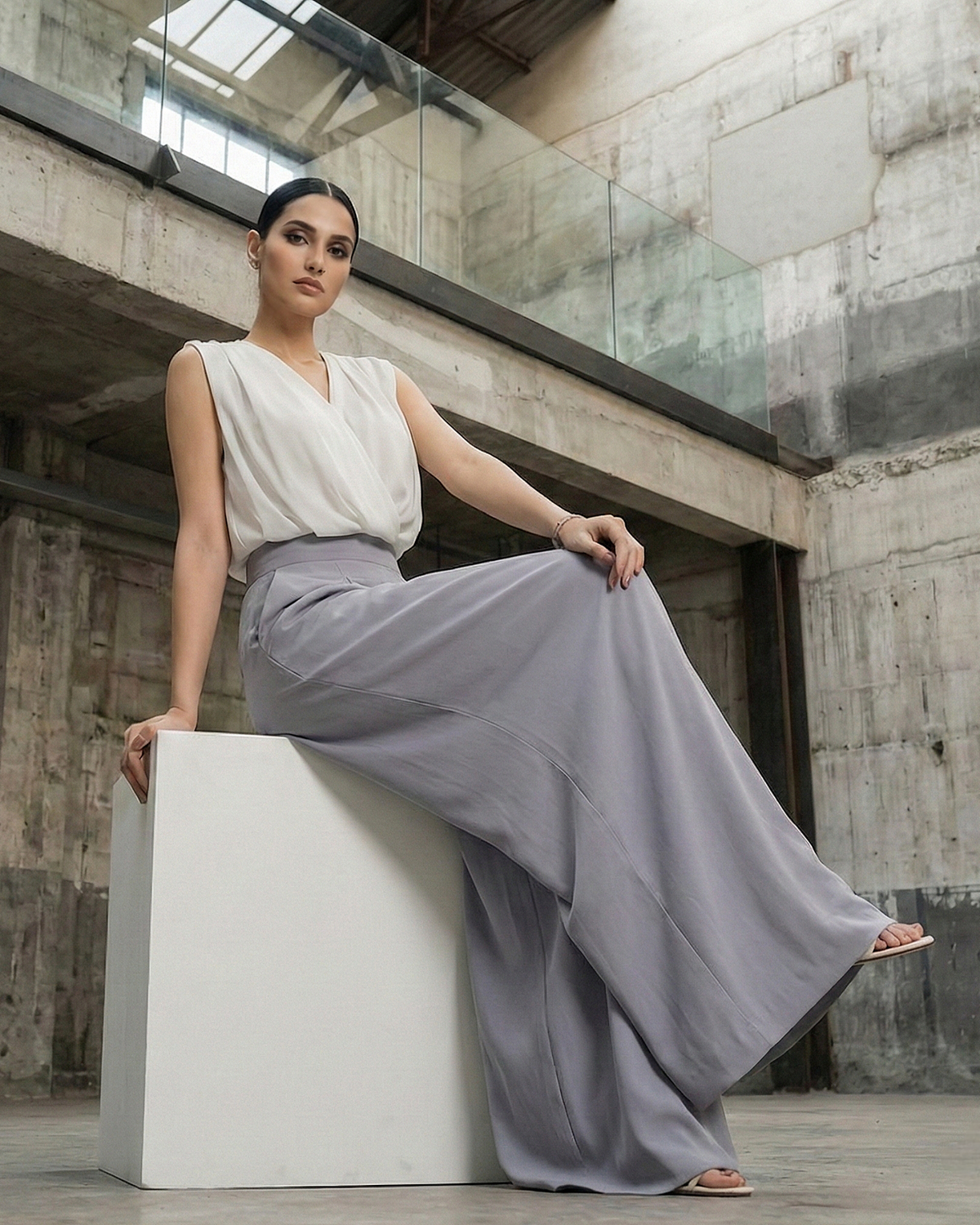 Woman in a white top and gray pants sitting on a white box in an industrial setting.